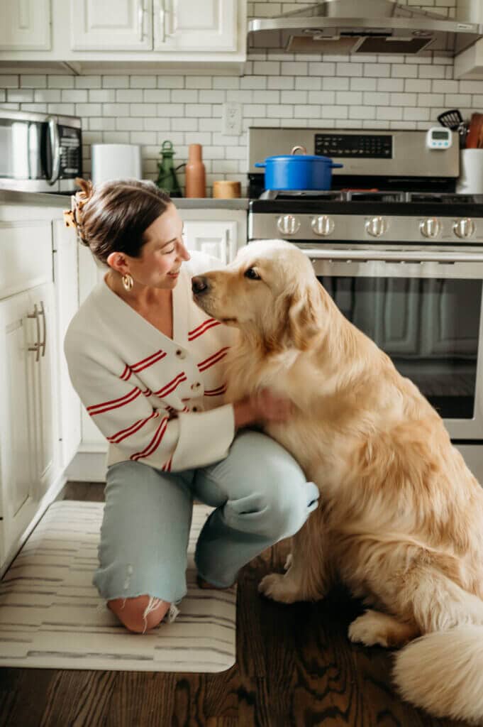 A woman kneels on a kitchen floor, smiling and petting a large golden retriever sitting beside her.