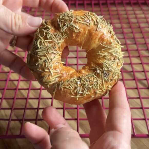 hands holding a rosemary sea salt greek yogurt bagel above a pink cooling rack